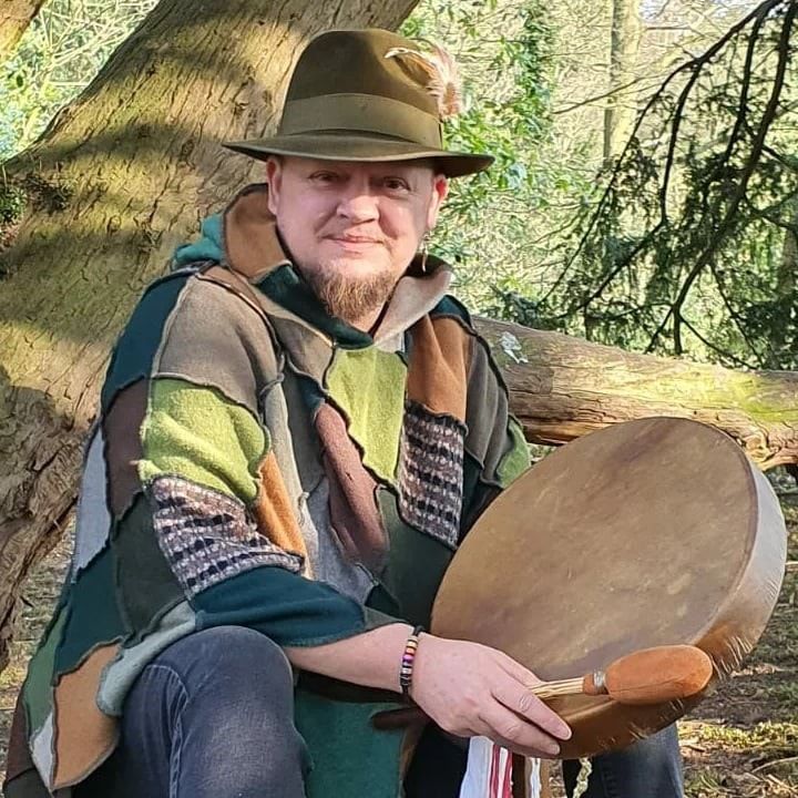 a picture in woodlands, green background, DK sat in a green hat and nature colours patchwork shawl, with his shamanic journey drum and a smile.